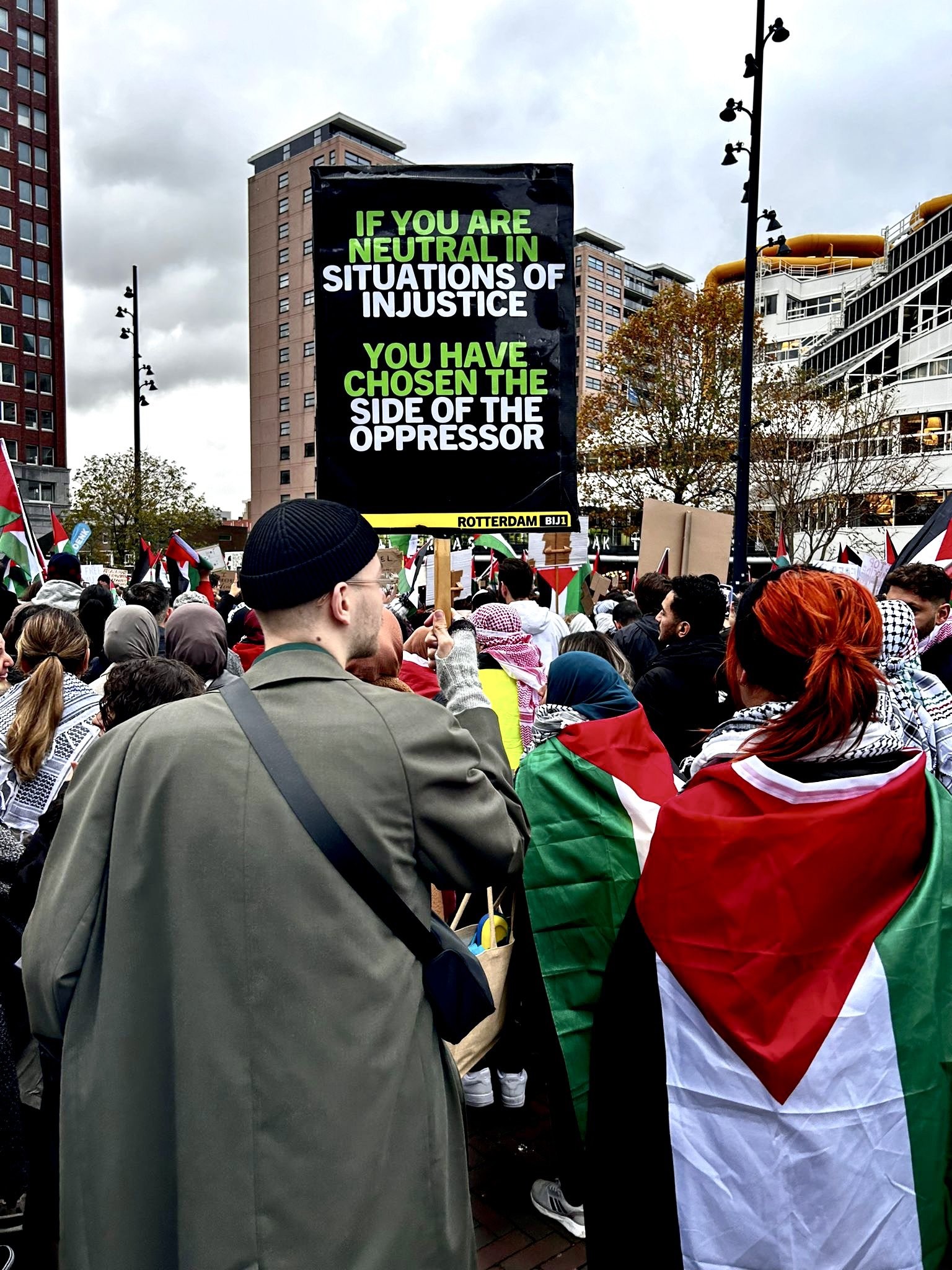 Foto van de demonstratie met een persoon die een bord van Rotterdam BIJ1 vasthoudt met de tekst: 'IF YOU ARE NEUTRAL IN SITUATIONS OF INJUSTICE, YOU HAVE CHOSEN THE SIDE OF THE OPPRESSOR'.