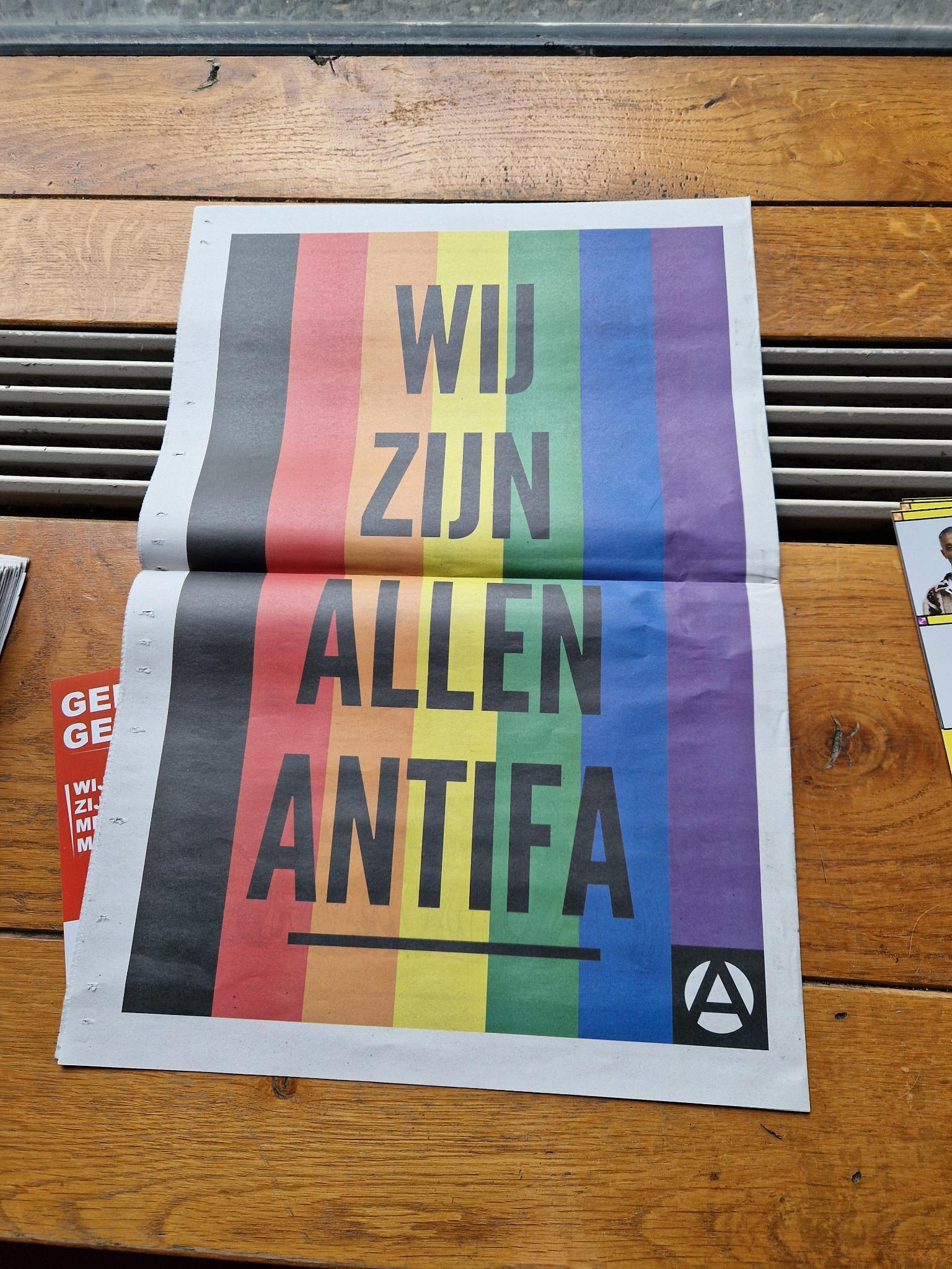 Antifaposter. Over de lengte lopen een aantal banen met de kleuren van de regenboog. Daaroverheen staat met zwarte letters "Wij zijn allen Antifa." Rechtsonder staat een klein logootje van een A in een wit cirkeltje op een zwart vierkantje.