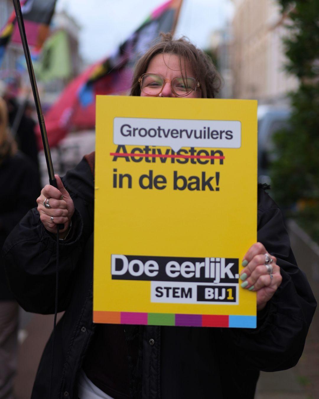 Een persoon met een boord met de tekst 'Grootvervuilers in de bak! Doe eerlijk stem BIJ1'