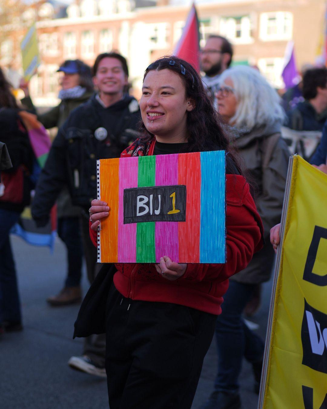 Een persoon met een bord in de BIJ1 kleuren en het woord BIJ1.