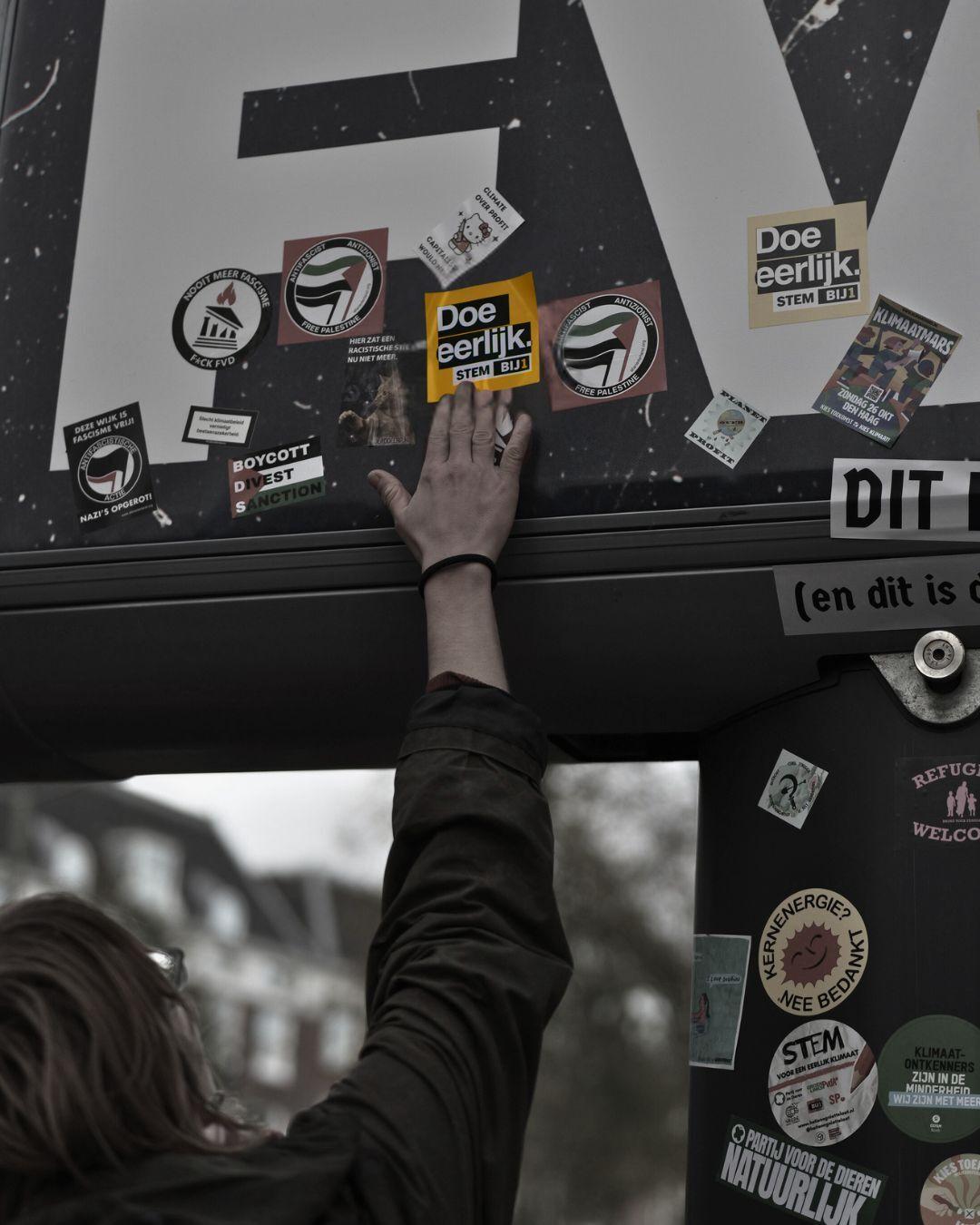 Iemand plakt en BIJ1-sticker op een mega groot verkiezingsbord van de FvD.