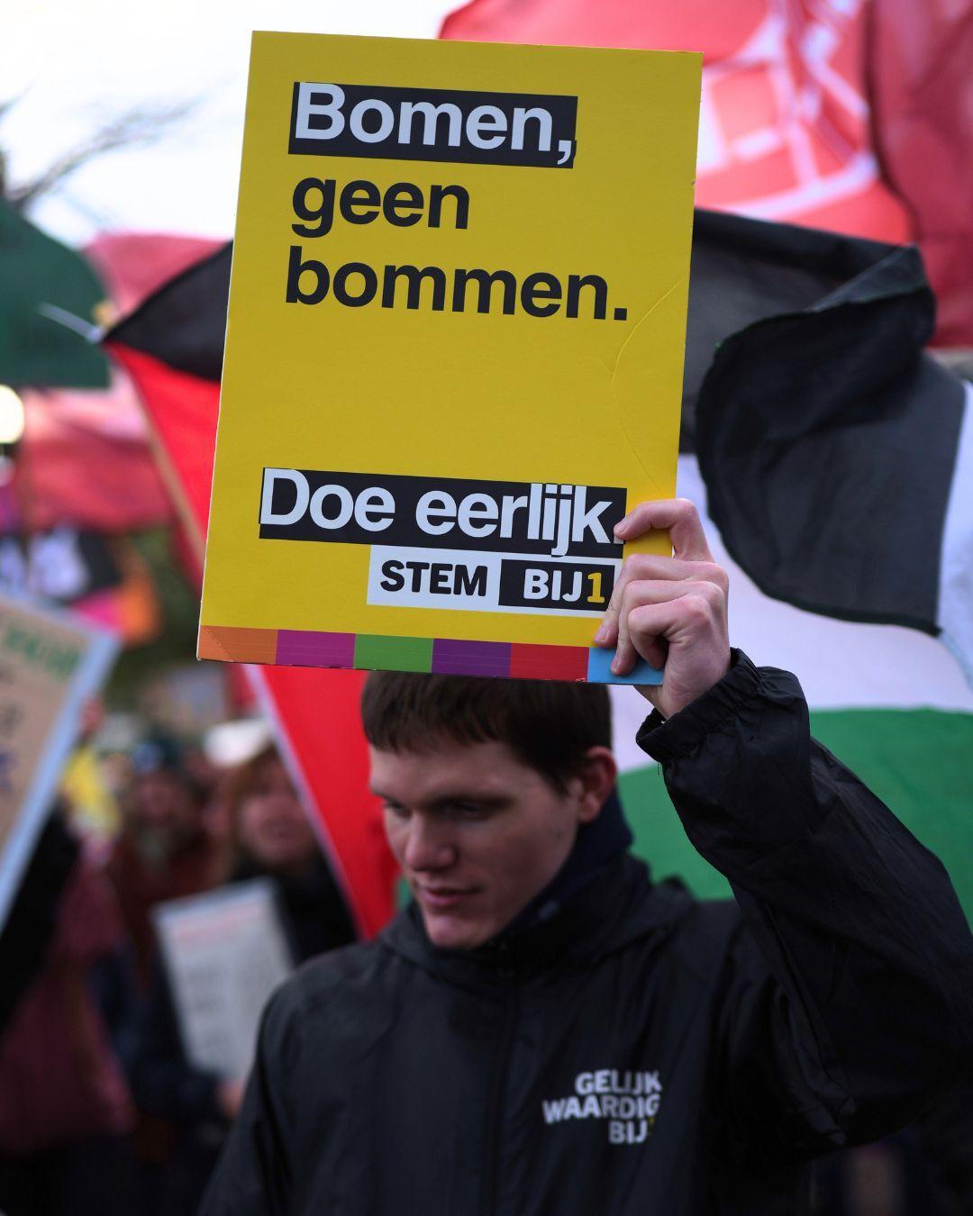 En persoon met en bord met de tekst 'Bomen, geen bommen. Doe eerlijk. STEM BIJ1'